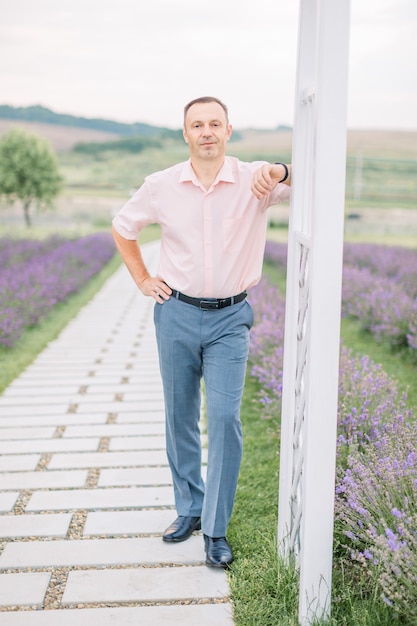 Homem bonito com camisa clara, aproveitando a caminhada, de pé no fundo do campo de lavanda, encostado no arco de madeira branco