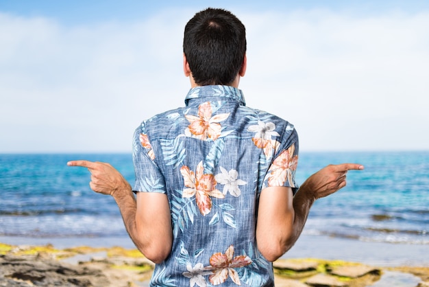 Homem bonito com camisa de flores apontando para os laterais tendo dúvidas na praia