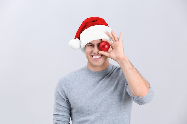 Homem bonito com chapéu de Natal segurando bugiganga brilhante sobre fundo claro