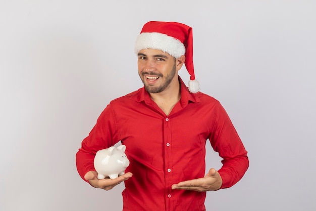 Homem bonito com chapéu de Natal segurando um cofrinho no fundo branco Conceito de economia de fim de ano