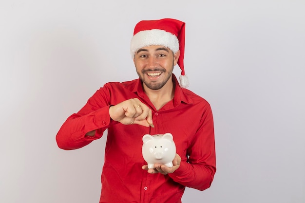 Homem bonito com chapéu de Natal segurando um cofrinho no fundo branco Conceito de economia de fim de ano