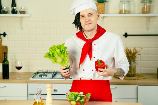 Homem bonito com dreadlocks no terno do chef com legumes frescos nas mãos em pé na cozinha Chef com salada de legumes cozidos em tigela transparente segurando alface pimenta vermelha Comida saudável