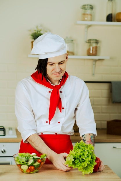 Homem bonito com dreadlocks no terno do chef com legumes frescos nas mãos em pé na cozinha Chef com salada de legumes cozidos em tigela transparente segurando alface pimenta vermelha Comida saudável