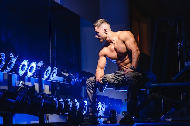 Homem bonito, com grandes músculos, posando para a câmera no ginásio. Fundo preto e azul. Retrato de um fisiculturista. Fechar-se.