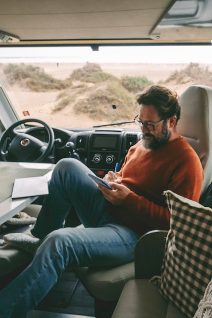 Foto homem bonito com óculos sentado dentro do camper escrevendo e fazendo anotações no caderno sorrindo acampamento atividade ao ar livre viagem de estrada conceito de natureza