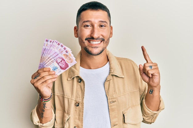 Homem bonito com tatuagens segurando notas de 50 pesos mexicanos sorrindo com uma ideia ou pergunta apontando o dedo com cara feliz número um