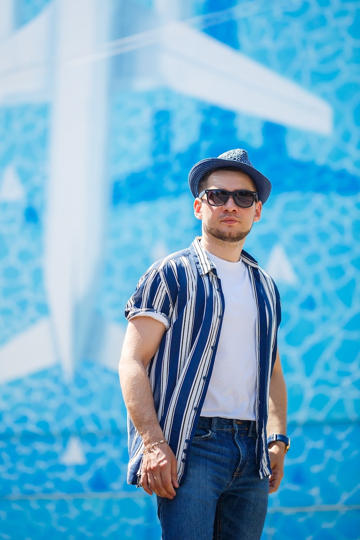 Foto homem bonito com uma barba em uma camiseta e uma camisa com um chapéu ficar sobre um fundo azul. cara de óculos escuros