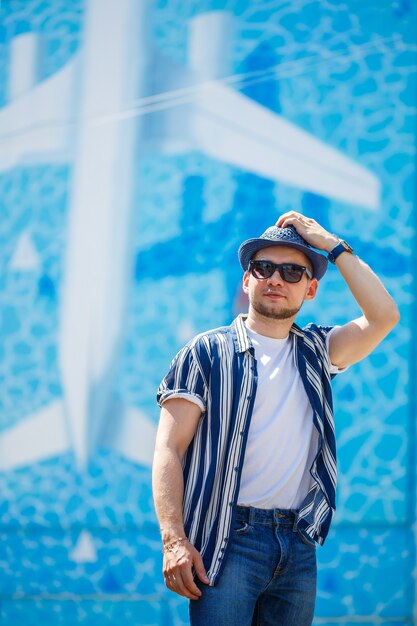 Homem bonito com uma barba em uma camiseta e uma camisa com um chapéu ficar sobre um fundo azul. Cara de óculos escuros