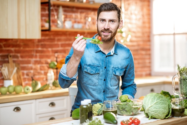 Foto homem bonito comendo salada saudável sentado à mesa cheia de ingredientes verdes na cozinha de casa