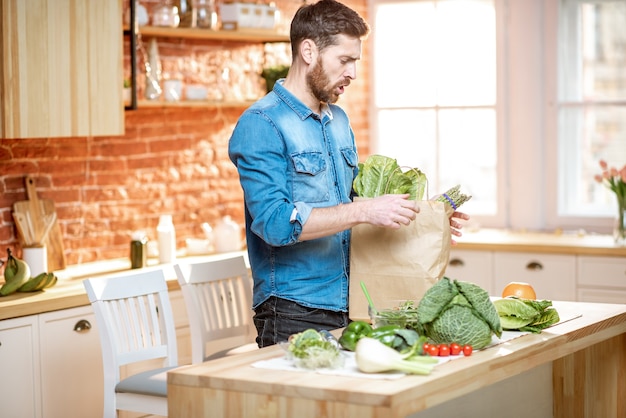 Homem bonito de camisa azul desempacotando comida saudável da sacola de compras na cozinha