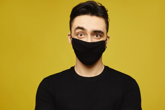 Homem bonito de camiseta preta e preta máscara protetora isolada em fundo amarelo. Doença sazonal e conceito de gripe sazonal. Conceito de estilo de vida em saúde