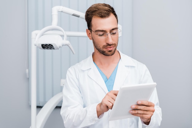 homem bonito de casaco branco e óculos usando tablet digital na clínica