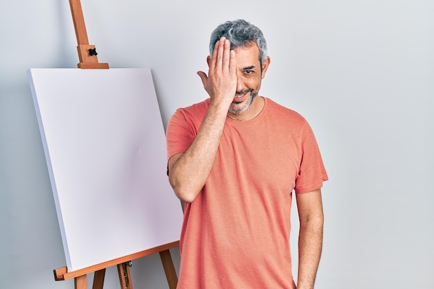 Homem bonito de meia-idade, com cabelos grisalhos, parado ao lado do cavalete do pintor, cobrindo um olho com a mão, sorriso confiante no rosto e emoção surpresa