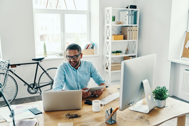 Homem bonito de meia-idade em roupa casual inteligente usando tablet digital e sorrindo enquanto está sentado no escritório