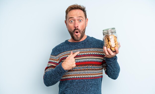 Homem bonito de meia-idade parecendo chocado e surpreso com a boca aberta, apontando para si mesmo. garrafa de biscoitos