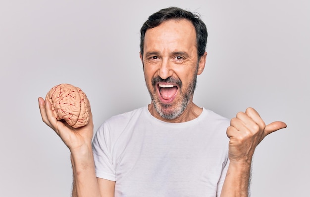 Homem bonito de meia-idade segurando cérebro de plástico sobre fundo branco isolado apontando o polegar para o lado sorrindo feliz com a boca aberta