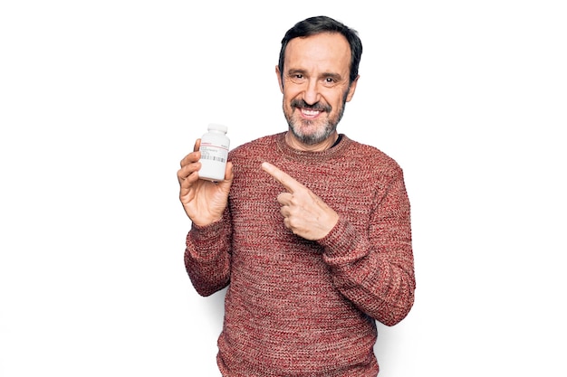 Homem bonito de meia-idade segurando o frasco de comprimidos de remédio sobre fundo branco isolado sorrindo feliz apontando com a mão e o dedo
