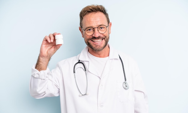 Homem bonito de meia-idade, sorrindo alegremente com uma mão no quadril e confiante. médico com frasco de comprimidos