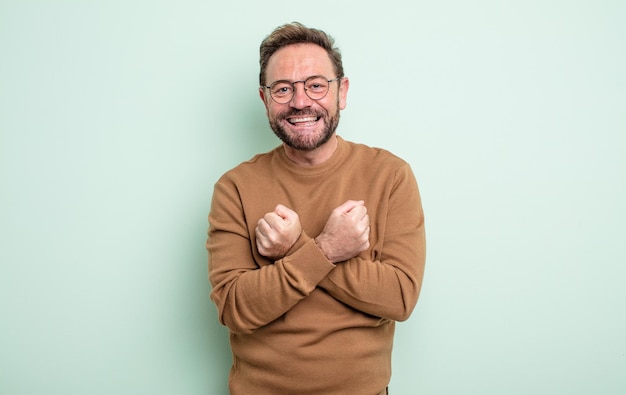 Homem bonito de meia-idade sorrindo alegremente e comemorando, com os punhos cerrados e os braços cruzados, sentindo-se feliz e positivo