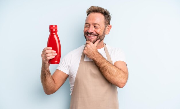 Homem bonito de meia-idade, sorrindo com uma expressão feliz e confiante com a mão no queixo. conceito de ketchup