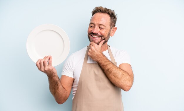 Homem bonito de meia-idade, sorrindo com uma expressão feliz e confiante com a mão no queixo. conceito de prato vazio