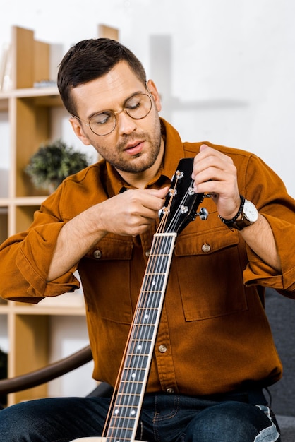 Foto homem bonito de óculos a afinar guitarra acústica em casa.