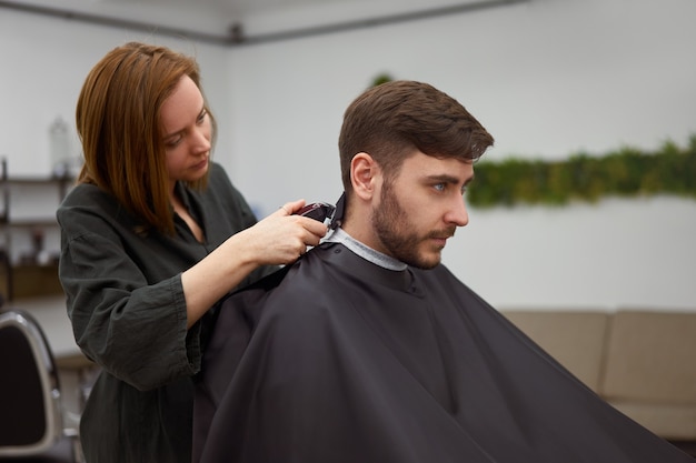 Homem bonito de olhos azuis sentado na barbearia. Mulher de cabeleireiro hairstylist cortando o cabelo dele. Barbeiro feminino.