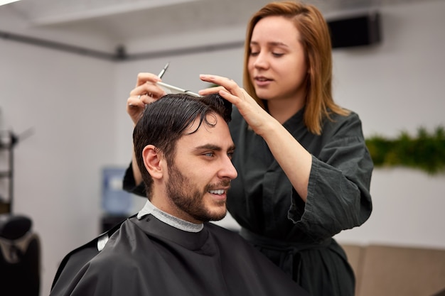 Homem bonito de olhos azuis sentado na barbearia. Mulher de cabeleireiro hairstylist cortando o cabelo dele. Barbeiro feminino.