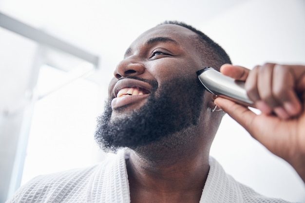 Homem bonito de pele escura com um sorriso no rosto enquanto faz procedimentos de beleza em seu banheiro