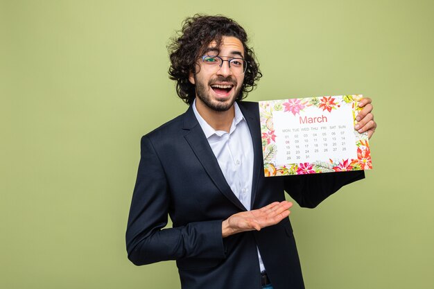 Homem bonito de terno segurando o calendário de papel do mês de março, apresentando o braço da mão, sorrindo, feliz e alegre, comemorando o dia internacional da mulher, 8 de março, em pé sobre fundo verde