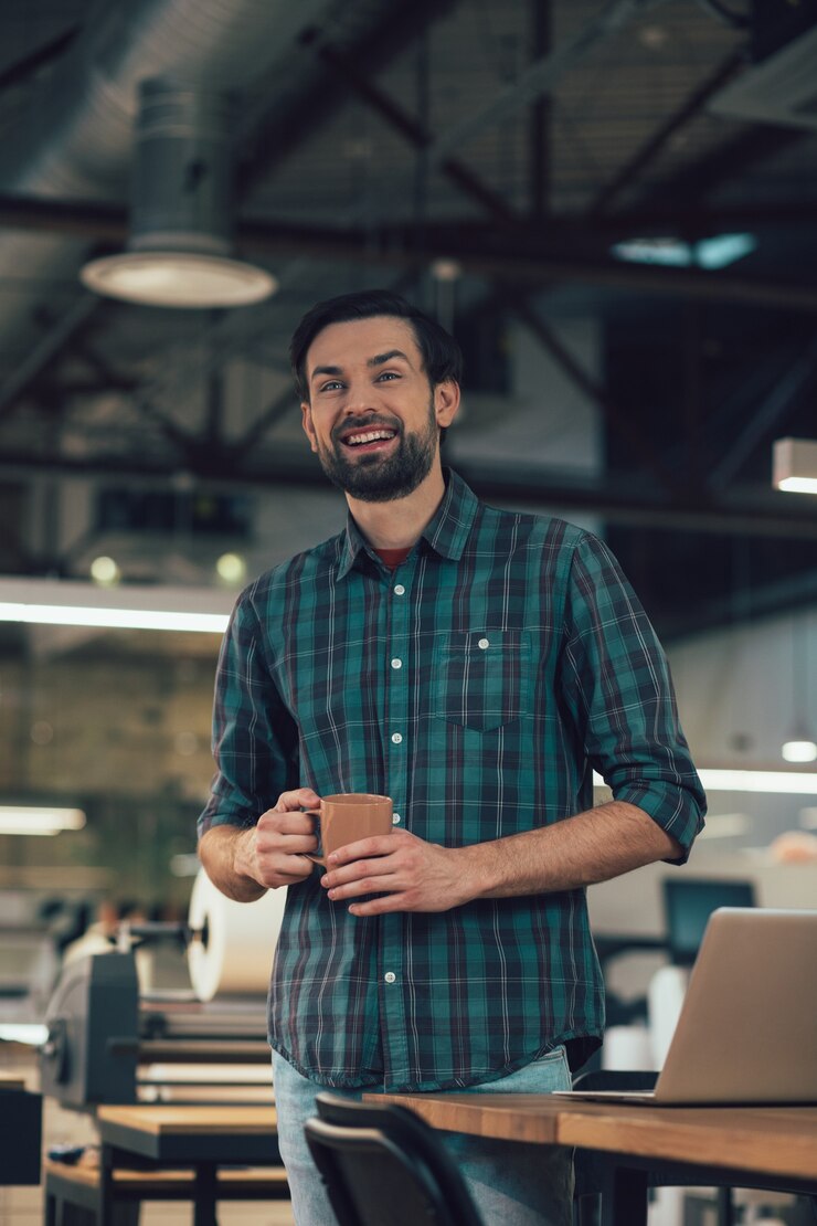 Foto homem bonito e alegre em pé perto da mesa e olhando para longe com um sorriso enquanto segura uma caneca