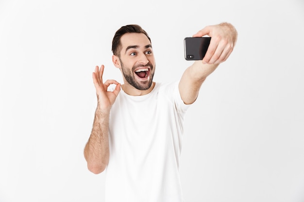 Homem bonito e alegre vestindo camiseta em branco, isolado na parede branca, ok