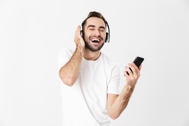 Homem bonito e alegre vestindo camiseta em branco, isolado na parede branca, ouvindo música com fones de ouvido, usando telefone celular