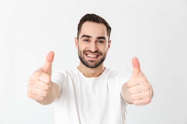 Homem bonito e alegre vestindo camiseta em branco, isolado na parede branca, polegar para cima