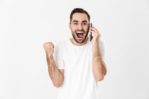 Homem bonito e alegre vestindo camiseta em branco, isolado na parede branca, usando telefone celular, comemorando o sucesso