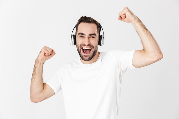 Homem bonito e alegre vestindo uma camiseta em branco, isolado na parede branca, ouvindo música com fones de ouvido