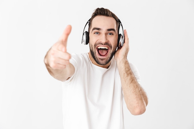 Homem bonito e alegre vestindo uma camiseta em branco, isolado na parede branca, ouvindo música com fones de ouvido