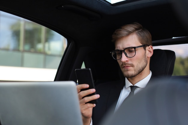 Homem bonito e confiante em terno completo, olhando para seu telefone inteligente enquanto está sentado no carro e usando o laptop.