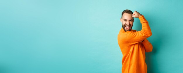 Homem bonito e confiante exibindo seus músculos flexionando o bíceps, sentindo-se forte em pé sobre o fundo azul claro