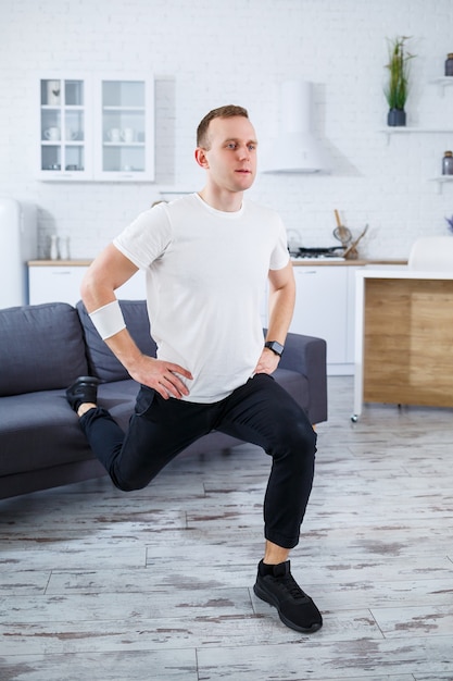 Homem bonito e desportivo com uma camiseta fazendo agachamentos em casa em seu apartamento. Fitness em casa. Estilo de vida saudável