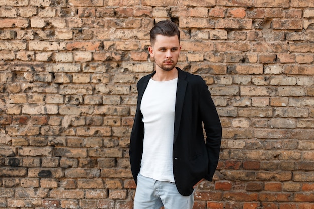 Homem bonito e elegante com uma jaqueta preta e camiseta branca posando perto de uma parede de tijolos