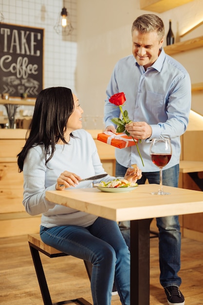 Homem bonito e feliz sorrindo e dando uma rosa e um presente para sua linda mulher de cabelos escuros alerta enquanto ela janta romântica