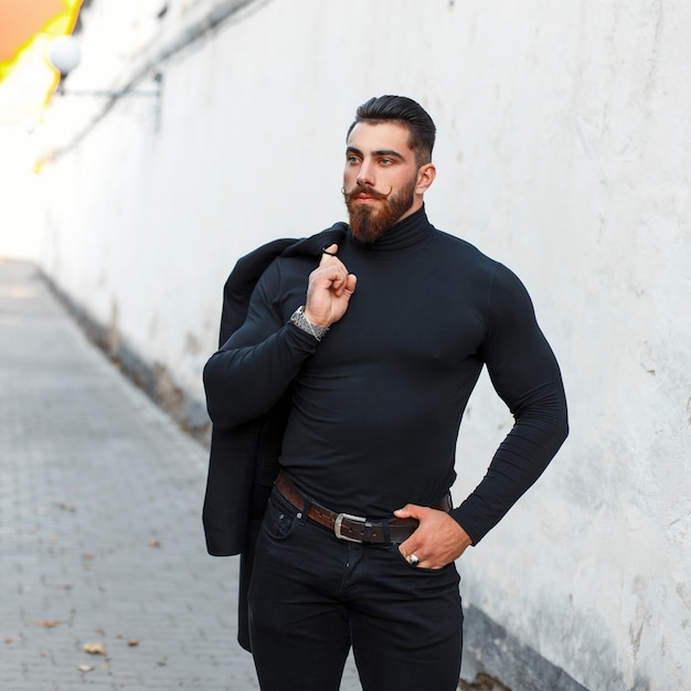 Homem bonito e forte com barba em roupas pretas elegantes posando na rua