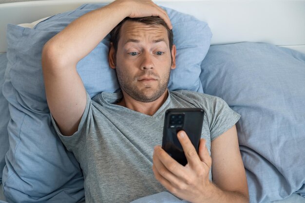 Foto homem bonito e impressionado lendo notícias no smartphone com expressão chateada na cama pela manhã.