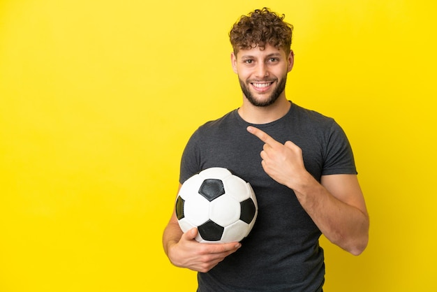 Homem bonito e jovem jogador de futebol isolado em fundo amarelo, apontando para o lado para apresentar um produto