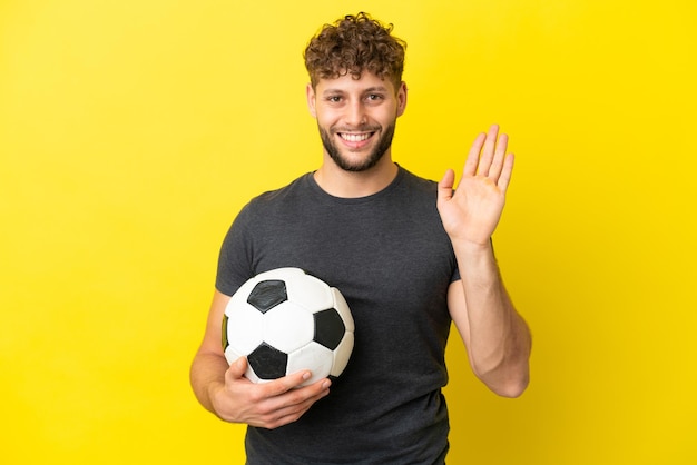 Homem bonito e jovem jogador de futebol isolado em um fundo amarelo, saudando com a mão e com uma expressão feliz