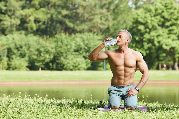Homem bonito e musculoso com abdômen na barriga sentado no tapete no parque e bebendo água da garrafa