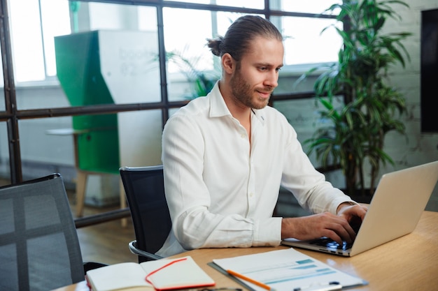 homem bonito e satisfeito com a barba por fazer trabalhando com um laptop enquanto está sentado à mesa em um escritório moderno