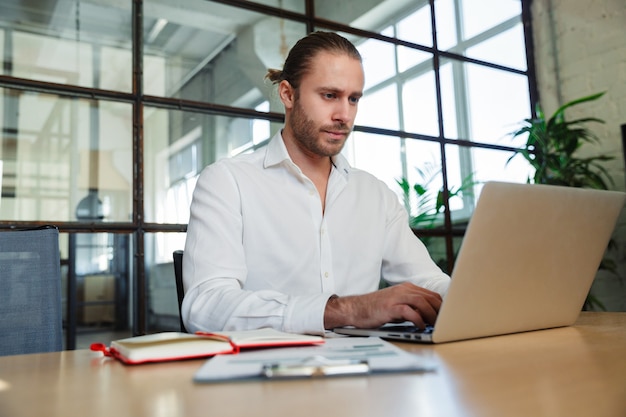 homem bonito e sério com barba por fazer trabalhando com laptop enquanto está sentado à mesa em um escritório moderno