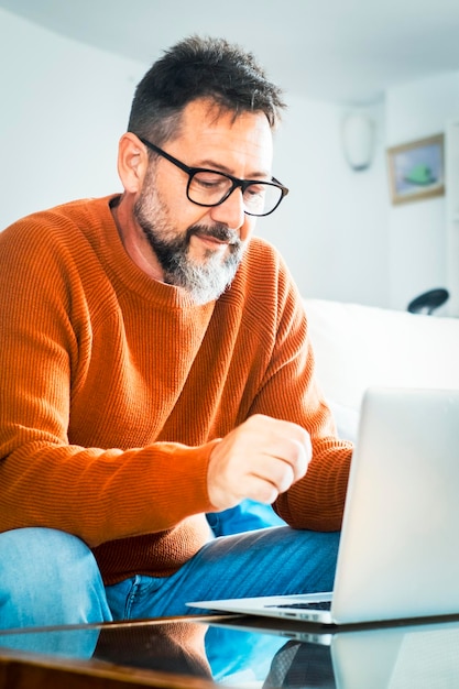 Homem bonito e sério senta-se no sofá com o computador em torno passa o tempo em casa navegando na web usando informações tecnologia moderna fazer pedidos na internet freelancing teletrabalho novo software pequeno negócio on-line